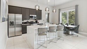 A modern kitchen with stainless steel appliances, a central island with bar stools, and a dining area with a large window. - Skyline Luxe Retreat  Loft, Pool Table & Rooftop Balcony Near Disney (Kissimmee)