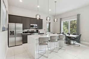 A modern kitchen with stainless steel appliances, a central island with bar stools, and a dining area with a large window. - Skyline Luxe Retreat  Loft, Pool Table & Rooftop Balcony Near Disney (Kissimmee)