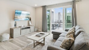 A living room with a sofa, a coffee table, a TV, and a sliding glass door leading to an outdoor area. - Skyline Luxe Retreat  Loft, Pool Table & Rooftop Balcony Near Disney (Kissimmee)