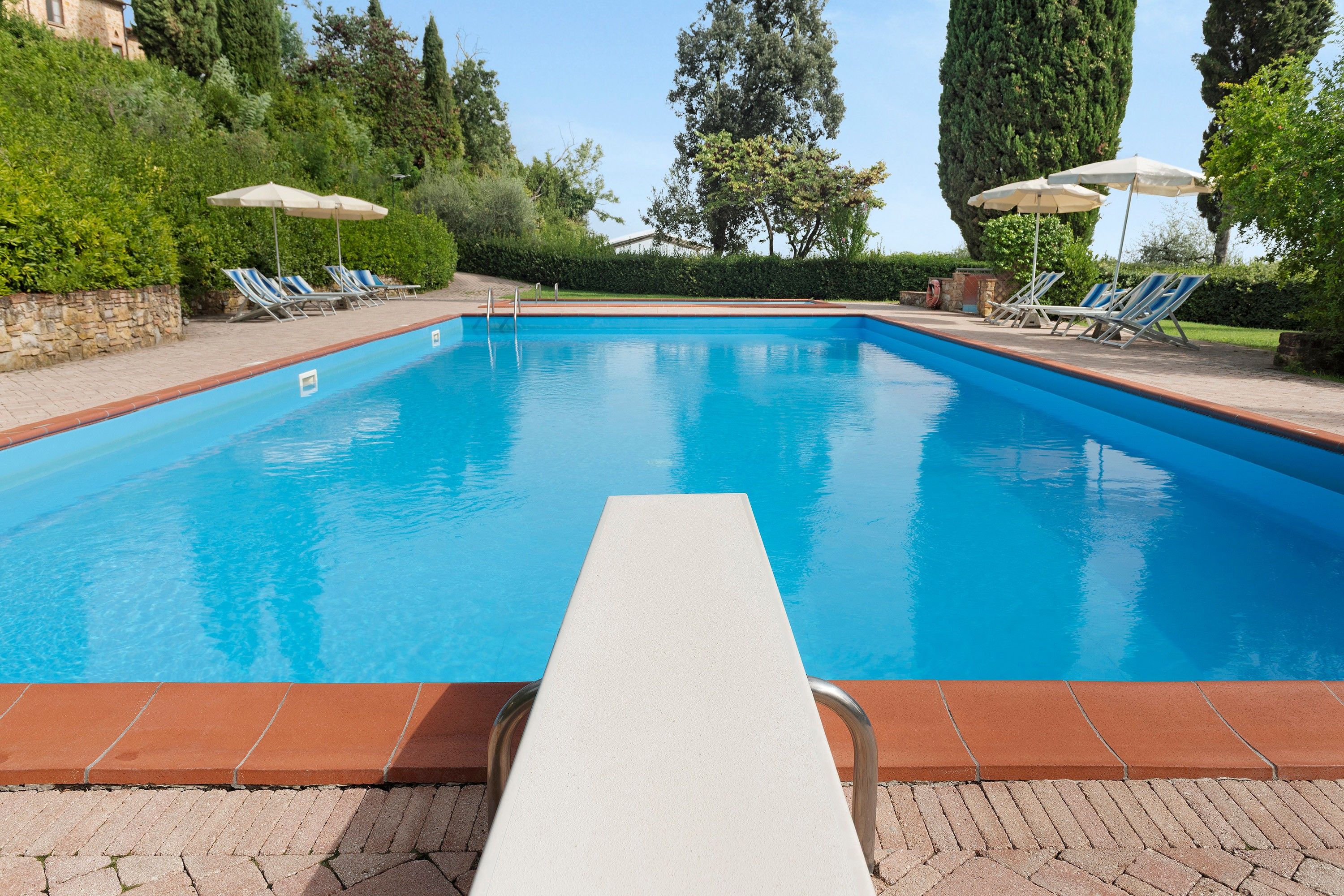 Una piscina rectangular amb vora de rajola, envoltada de gandules i ombrel·les, i un camí de maó que hi condueix.