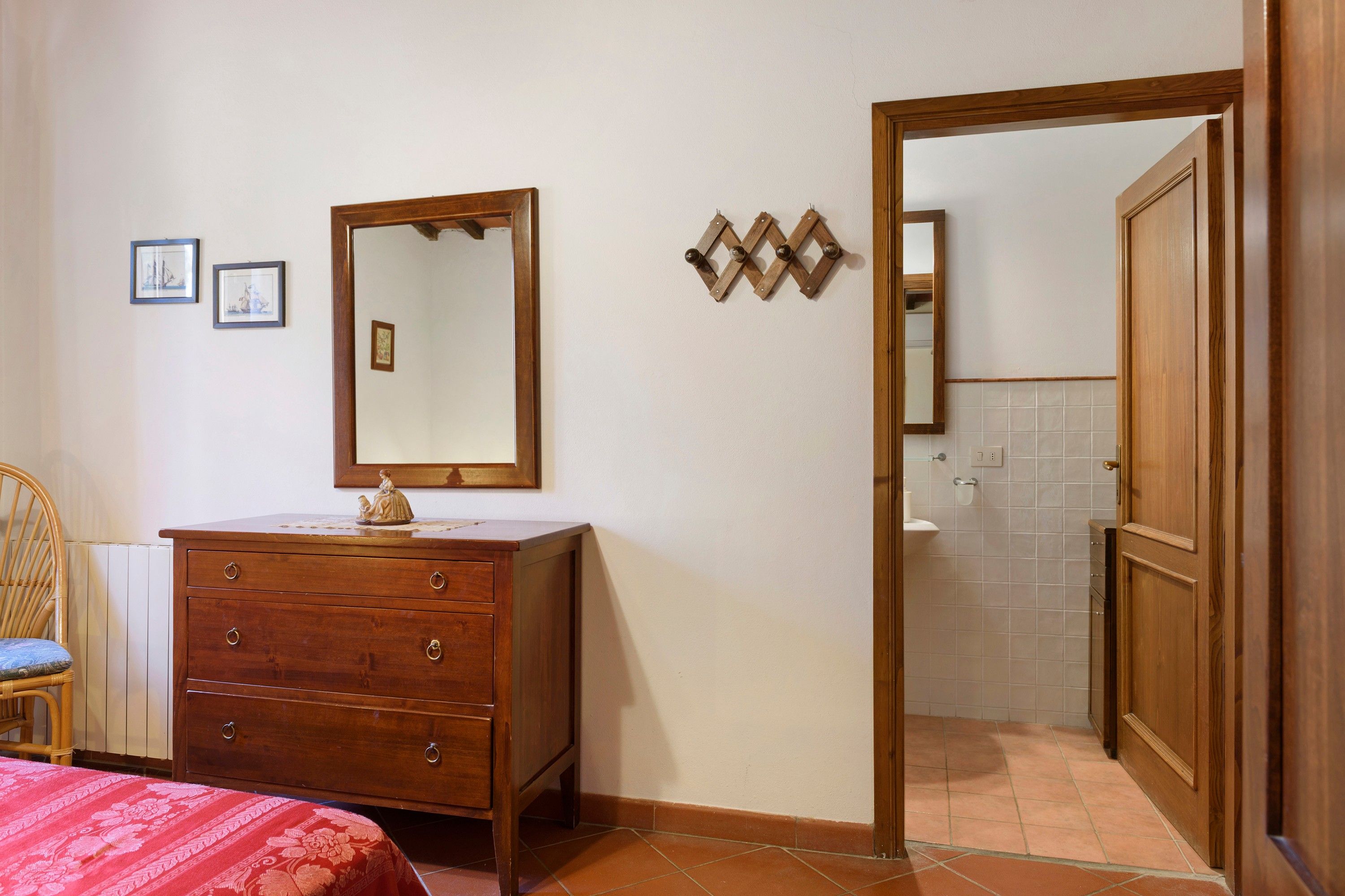 Een slaapkamer met een houten dressoir, een bed met een rode sprei, een stoel en een zichtbare badkamer door een open deur.