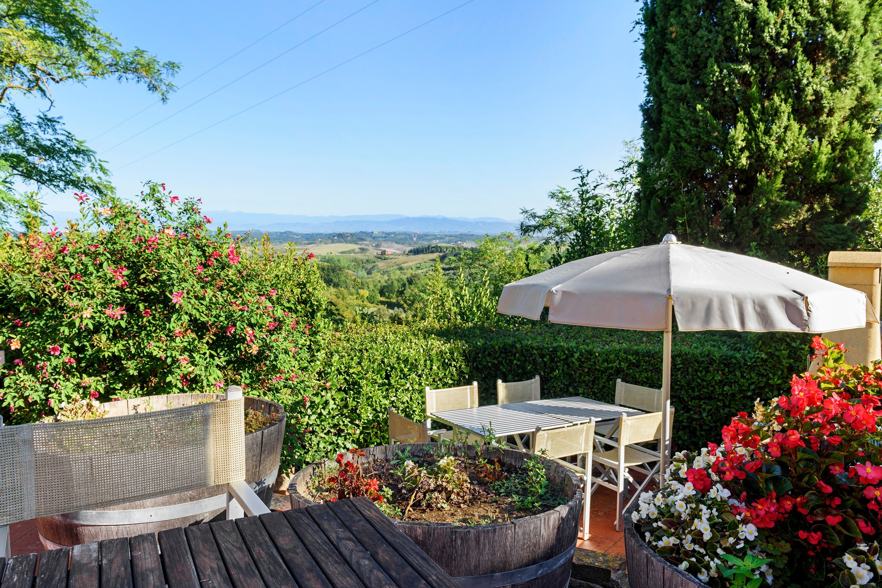 A patio with a table and chairs, surrounded by flowering plants and a view of the countryside.