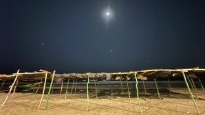 Vista nocturna de una playa con una luna brillante y estrellas, un primer plano arenoso y una estructura improvisada con postes verdes.