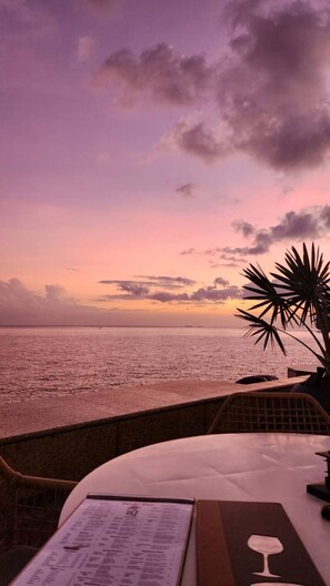 A seaside view with a menu on a table, palm trees, and a sunset. - The Nook at Maison Rouge: Classy, Cosy, Comfort (Port of Spain)
