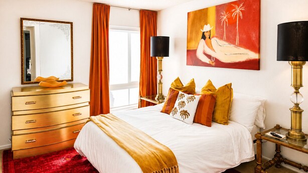 Una camera d'albergo con un letto, una cassettiera, uno specchio, un quadro e una lampada.