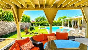 Zona relax all'aperto coperta, con tavolo in vetro, cuscini arancioni e vista su una piscina.
