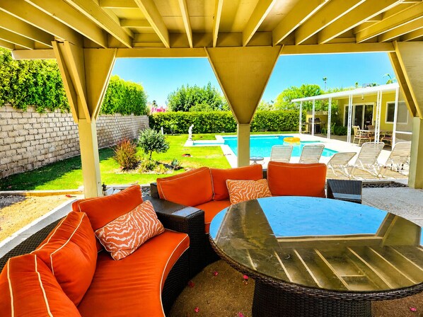 Un área de estar al aire libre cubierta, con una mesa de vidrio, cojines naranjas y vista a una piscina.