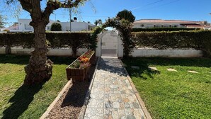 Un chemin menant à une entrée fermée par un portail, bordé de haies bien entretenues et d’un arbre.