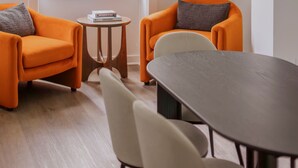Una habitación con una pared blanca curva, un ventanal grande, dos sillones anaranjados, una mesita redonda y una mesa de madera oscura.