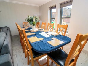 A dining area with a table set for six, surrounded by wooden chairs with black cushions. A vase with flowers is placed on the table. - Ledrishmore View (Alexandria)
