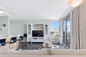 A modern living room with a TV, seating, a dining area, and a balcony with a city view. - Estero Beach & Tennis 908-b (Fort Myers Beach)