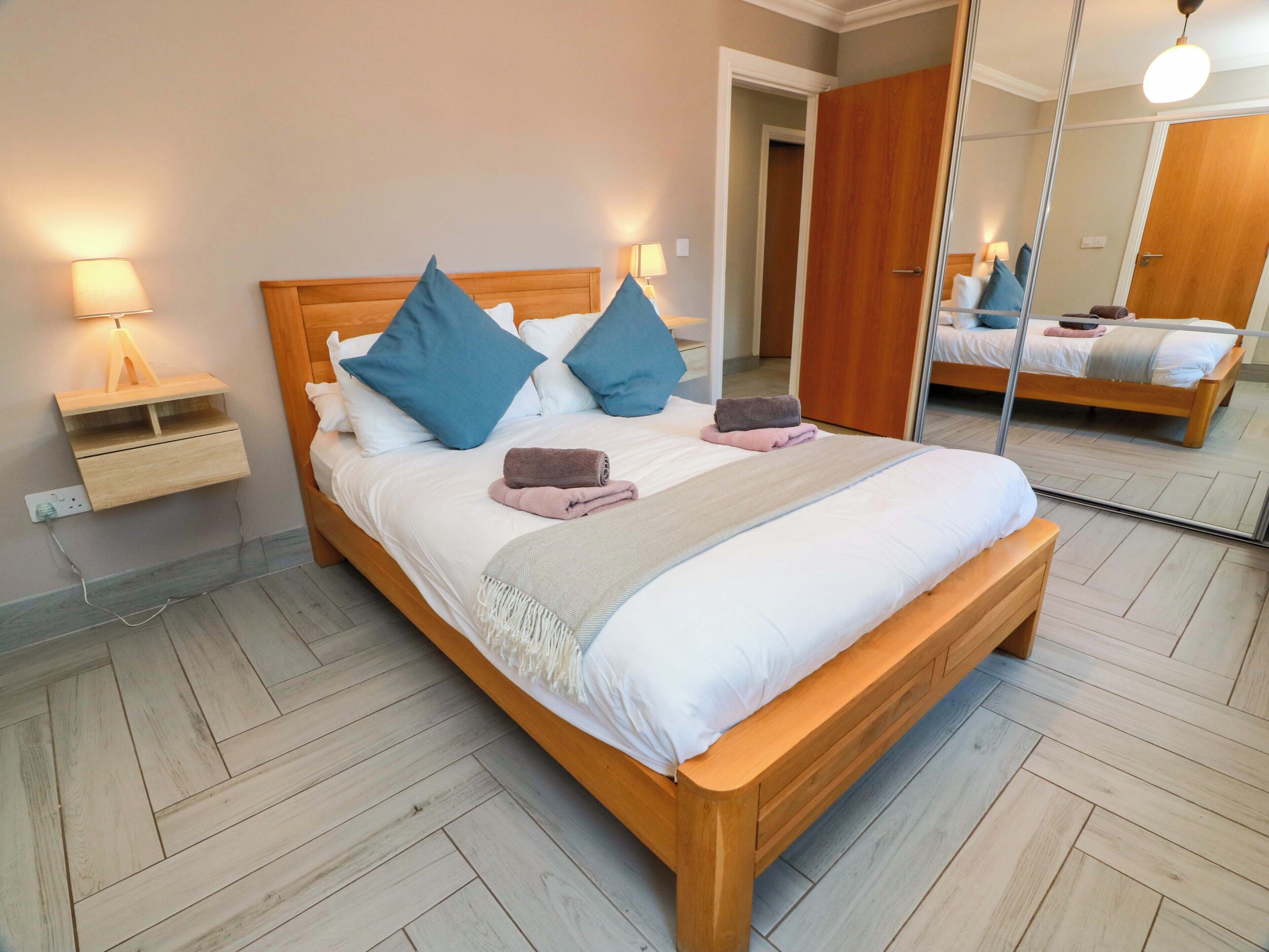 A neatly made bed with pillows and a blanket, a bedside table with a lamp, and a large mirror reflecting the room.