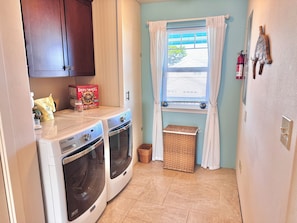 A laundry room with two front-loading washing machines, a drying rack, a wicker basket, and a window with blinds.