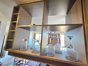 A wooden shelf with glassware, including wine glasses and a decanter, in a room with a window and a painting.