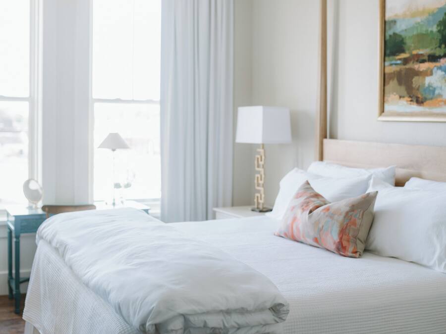 A neatly made bed with white linens, a bedside table with a lamp, and a painting on the wall.