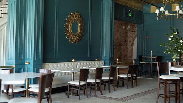 Interior de restaurante con paredes color turquesa, un banco acolchado blanco, sillas de madera y candelabros colgantes.