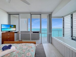 Ein Schlafzimmer mit Bett, Fernseher, Holzkommode, Balkon mit Meerblick und Fenster mit Jalousien.