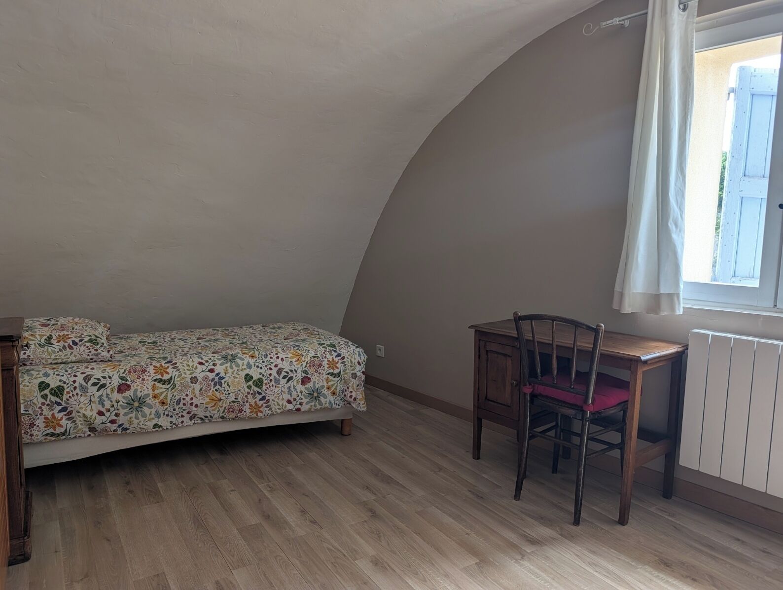 Une chambre à coucher avec un plafond incurvé, un lit recouvert d’une couvre-lit à motifs floraux, une chaise en bois et une fenêtre avec des rideaux.