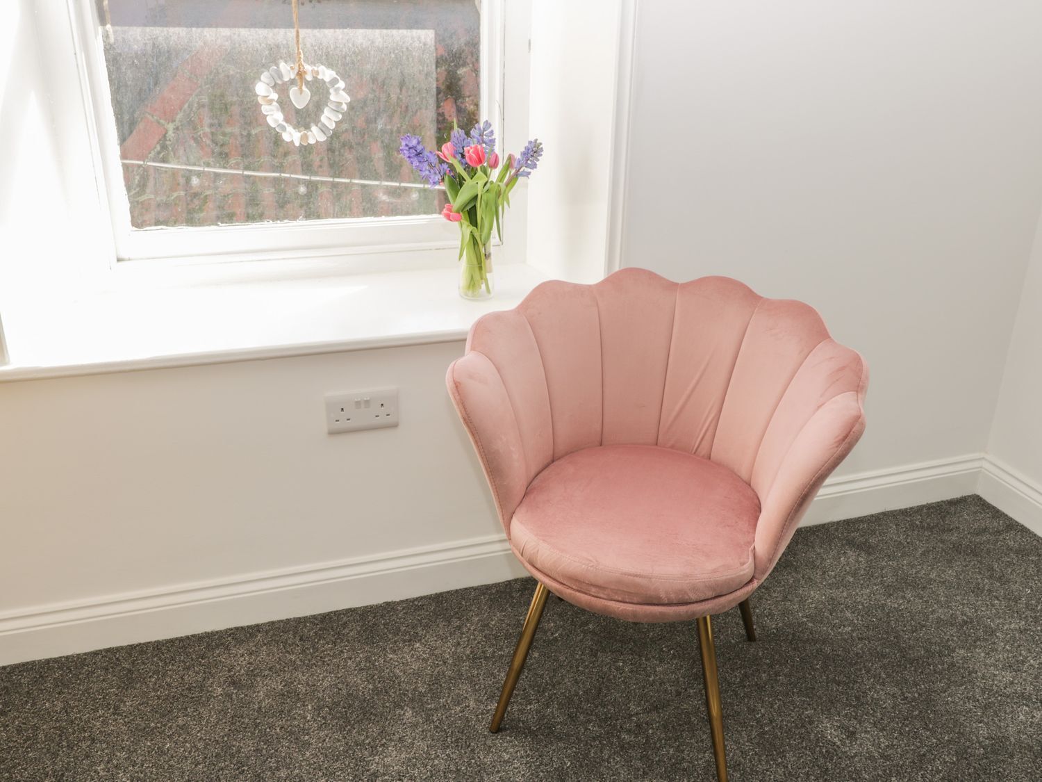 A pink chair with gold legs in a room with a window, carpeted floor, and a vase with flowers.