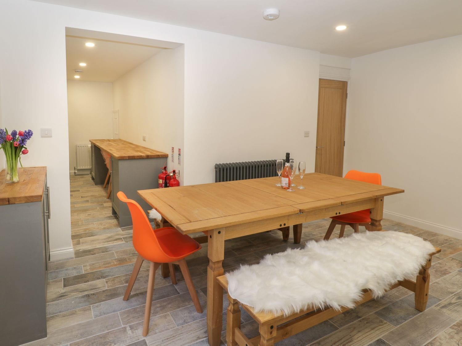 Een eethoek met een houten tafel, oranje stoelen, een zitbank van bont en een keuken op de achtergrond.