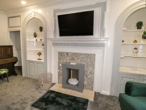 A living room with a fireplace, TV, shelves with plants, a piano, and a green sofa. - 47 Baxtergate (Whitby)