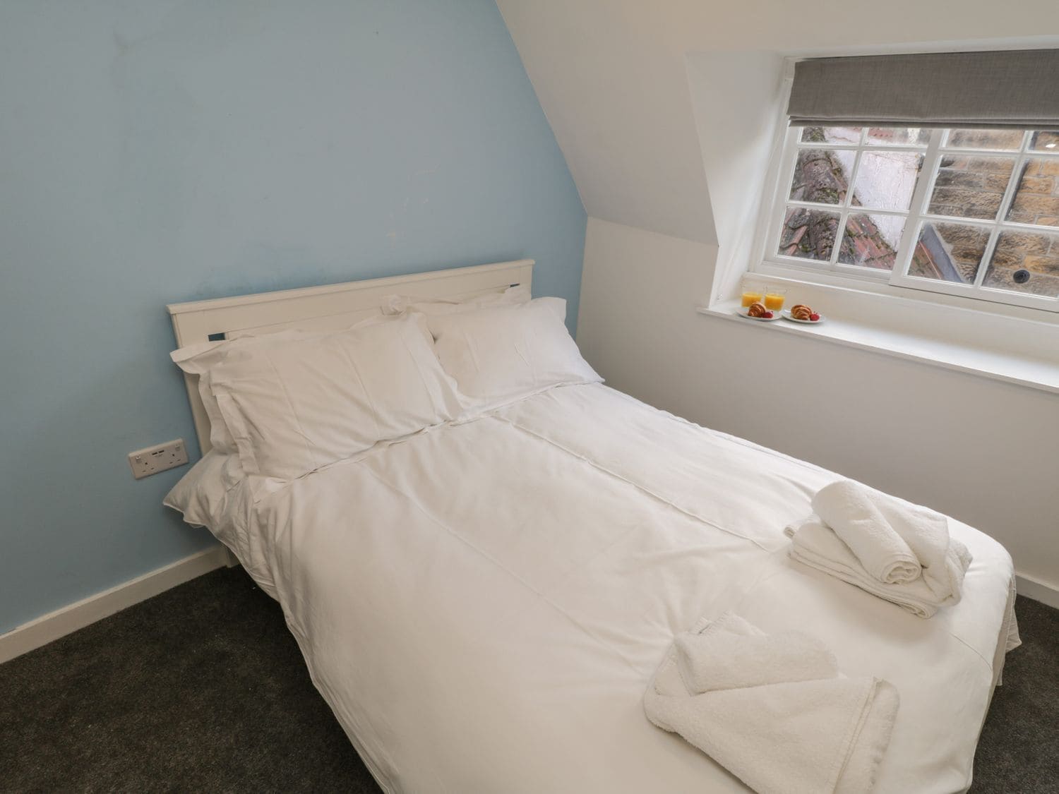 A single bed with white bedding, a window with a view of the outside, and a small shelf with items.