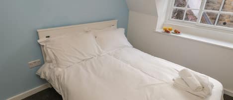 A single bed with white bedding, a window with a view of the outside, and a small shelf with items.