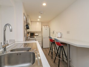 A modern kitchen with a stainless steel sink, a microwave, a toaster, and a refrigerator. There are two bar stools with patterned seats. - 47A Baxtergate (Whitby)