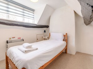 A single bed with white linens, a bedside table with a lamp, a radiator, and a window with blinds. - The Anchorage (Whitby)