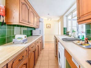 A kitchen with wooden cabinets, a microwave, a washing machine, and a sink with various items on the counter. - The Anchorage (Whitby)