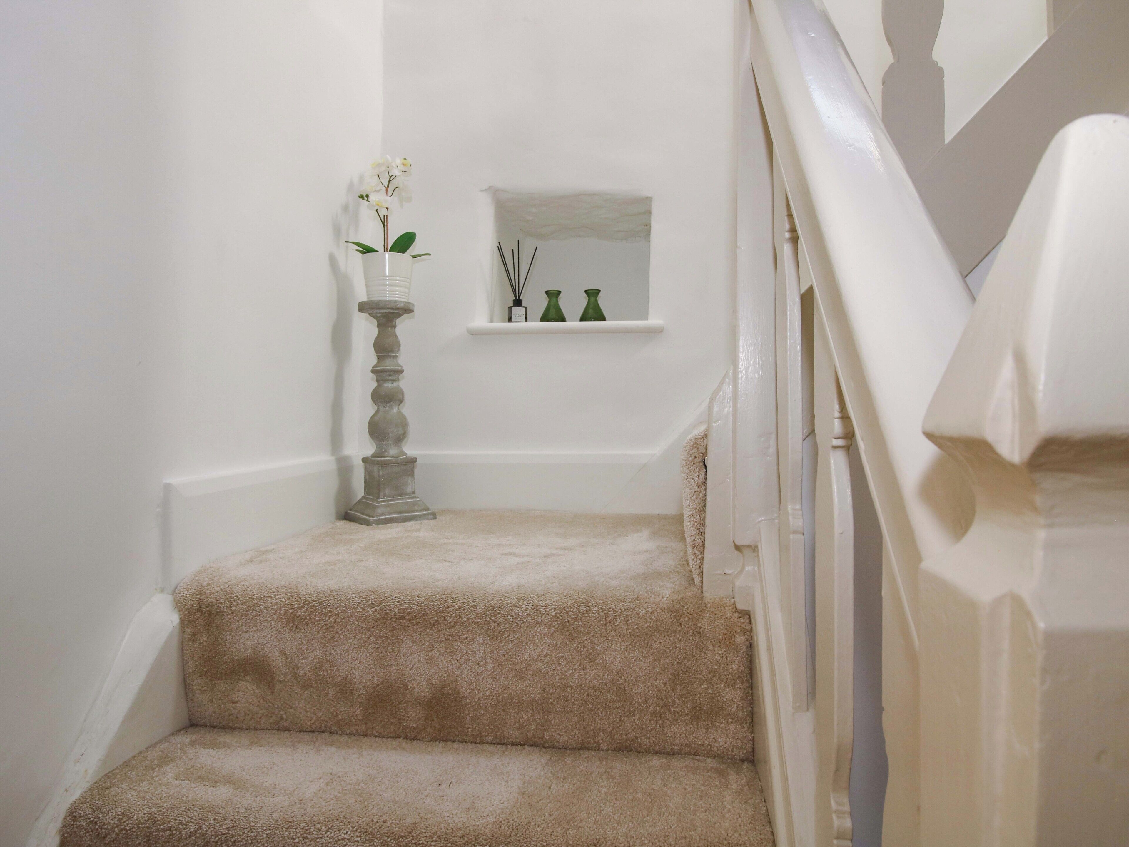 A carpeted staircase with a white wall, a small shelf with decorative items, and a tall vase with flowers.