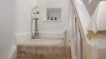 A carpeted staircase with a white wall, a small shelf with decorative items, and a tall vase with flowers.