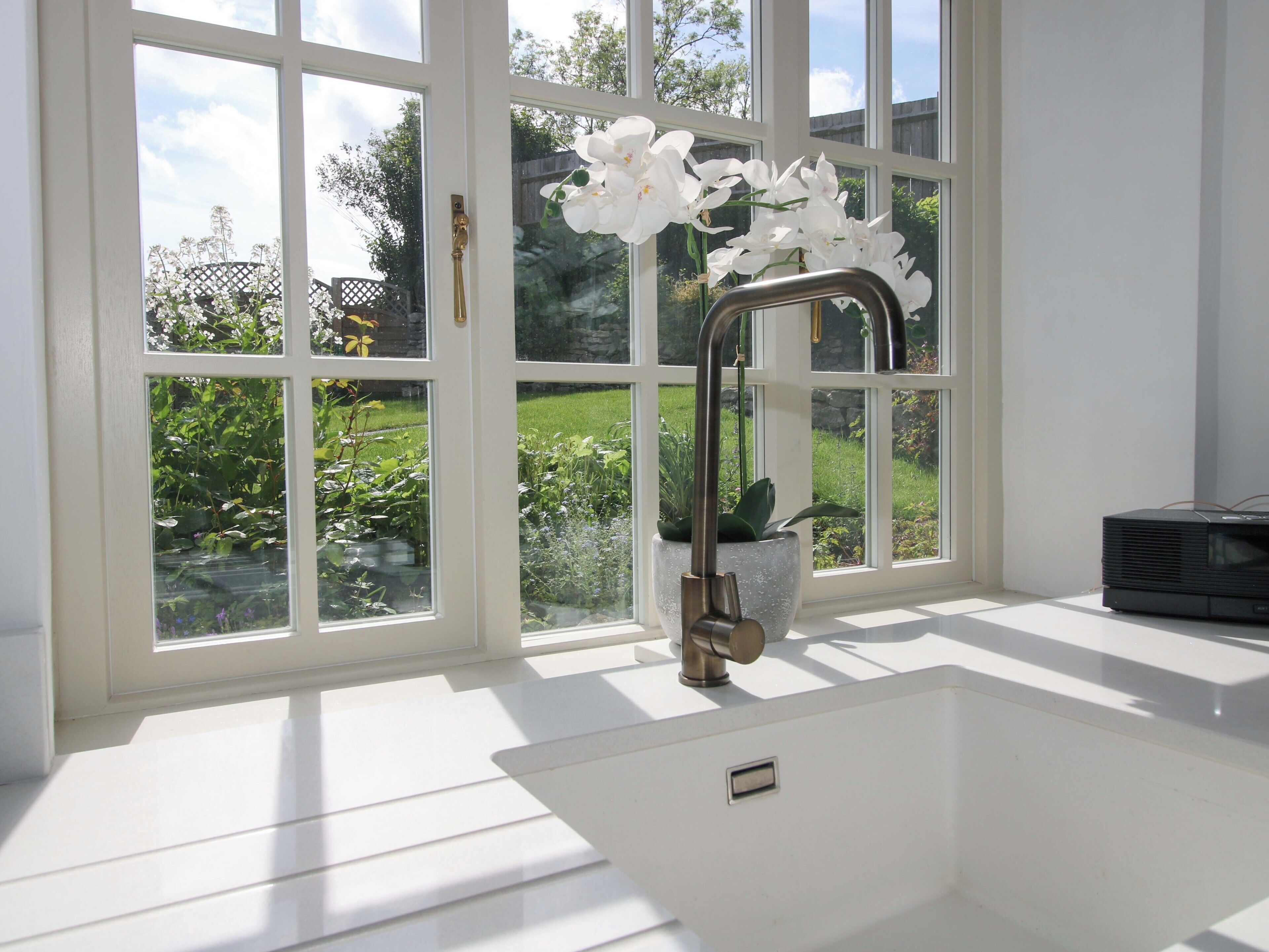 Casa de banho com lavatório branco, torneira moderna e vista para um jardim através de grandes janelas.