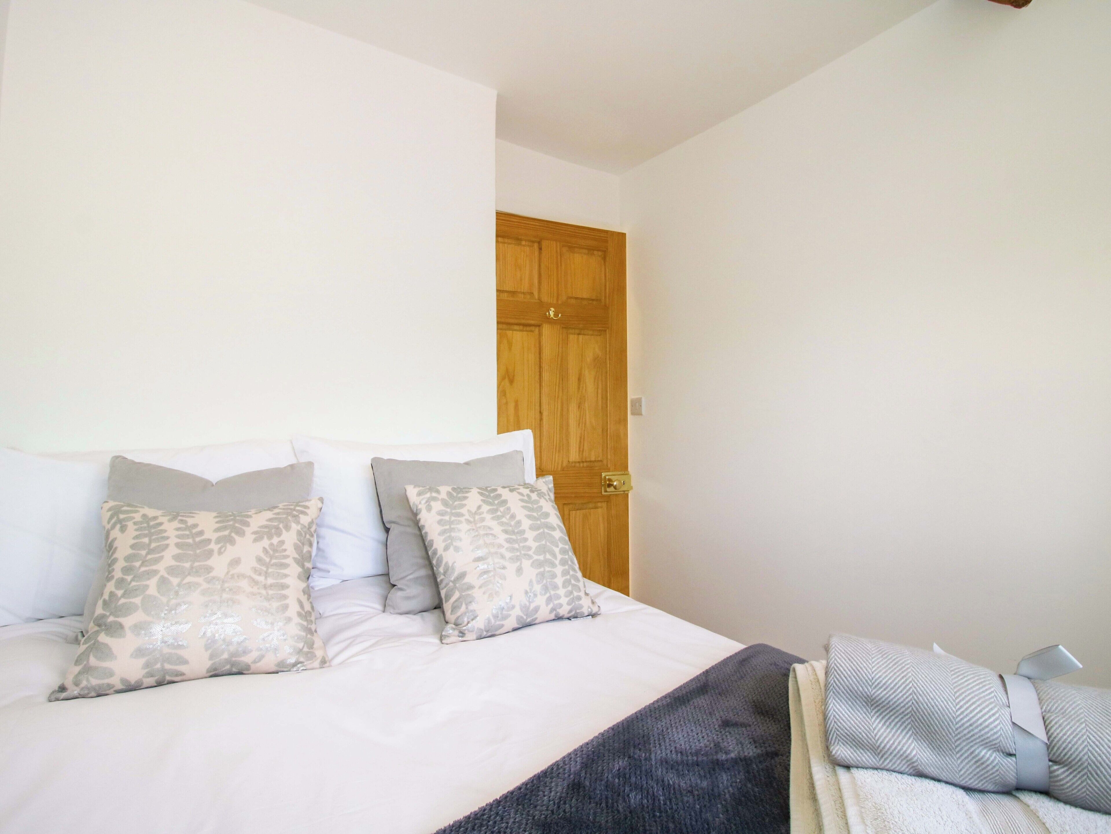 Quarto com cama, almofadas, manta, porta de madeira e uma pequena mesa com abajur.