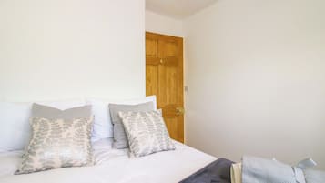 Quarto com cama, almofadas, manta, porta de madeira e uma pequena mesa com abajur.