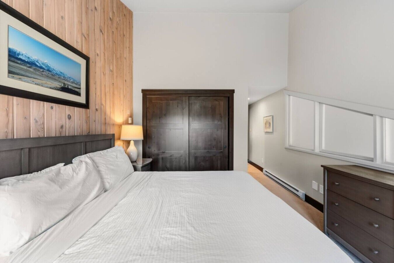 A hotel room with a bed, a wooden headboard, a door, a dresser, and a framed picture on the wall.