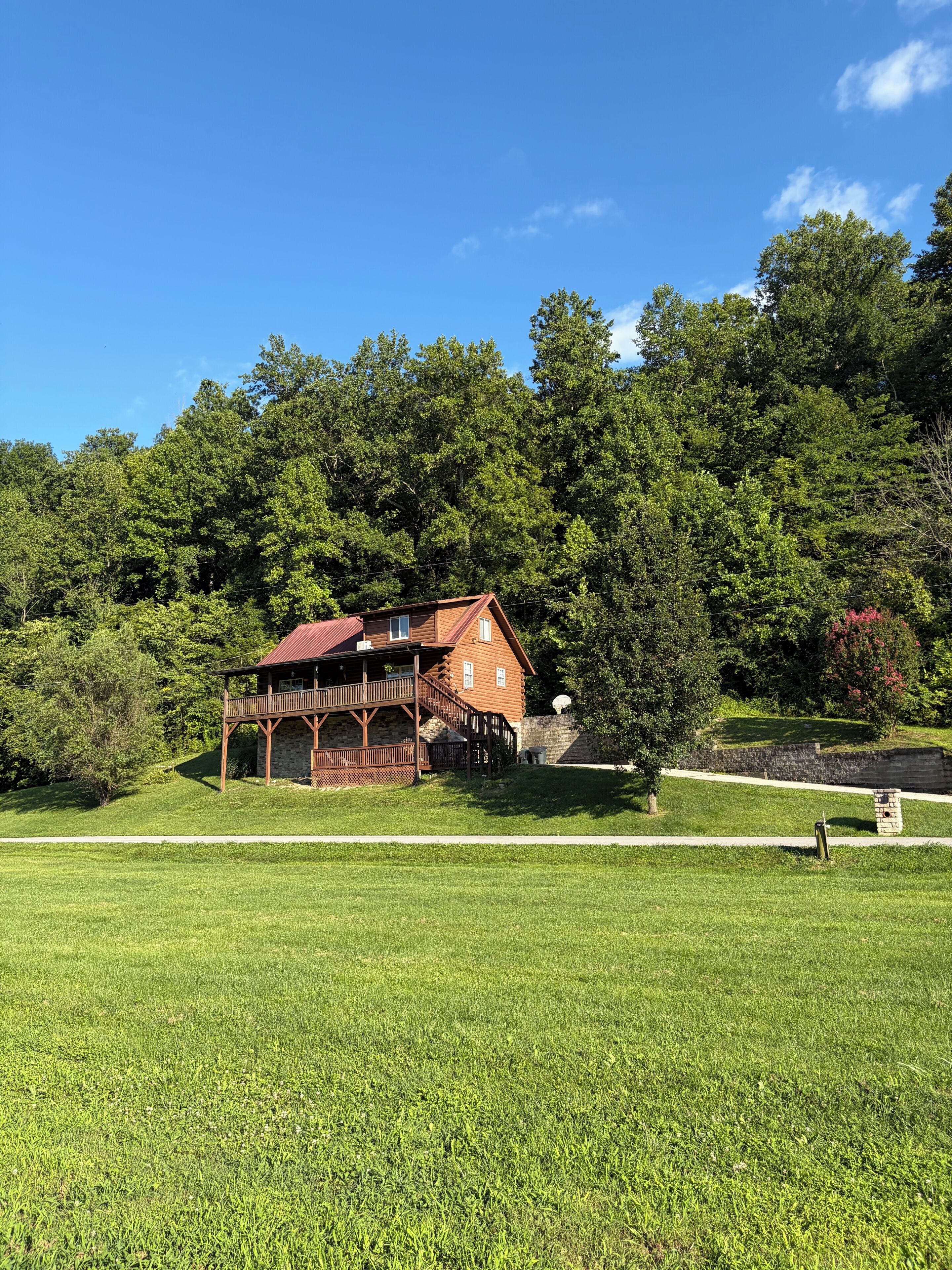 A wooden cabin with a porch and a stone wall, surrounded by a well-maintained lawn and dense forest.