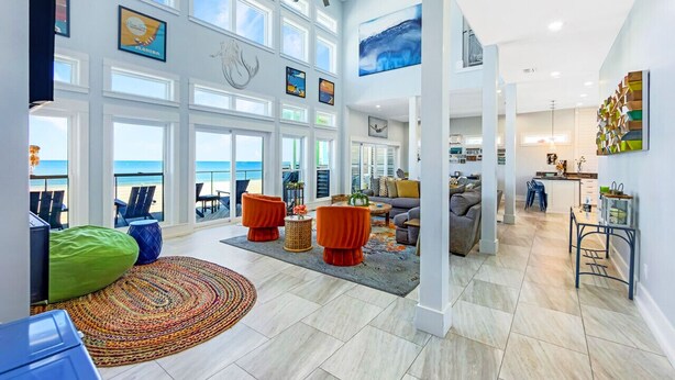 Een ruime woonkamer met een groot raam met uitzicht op het strand, een plafondventilator en een moderne keuken op de achtergrond.