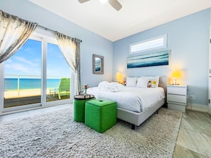 Ein Schlafzimmer mit einem Bett, einem Nachttisch, einer Lampe, einem grünen Sessel und Blick auf den Strand.