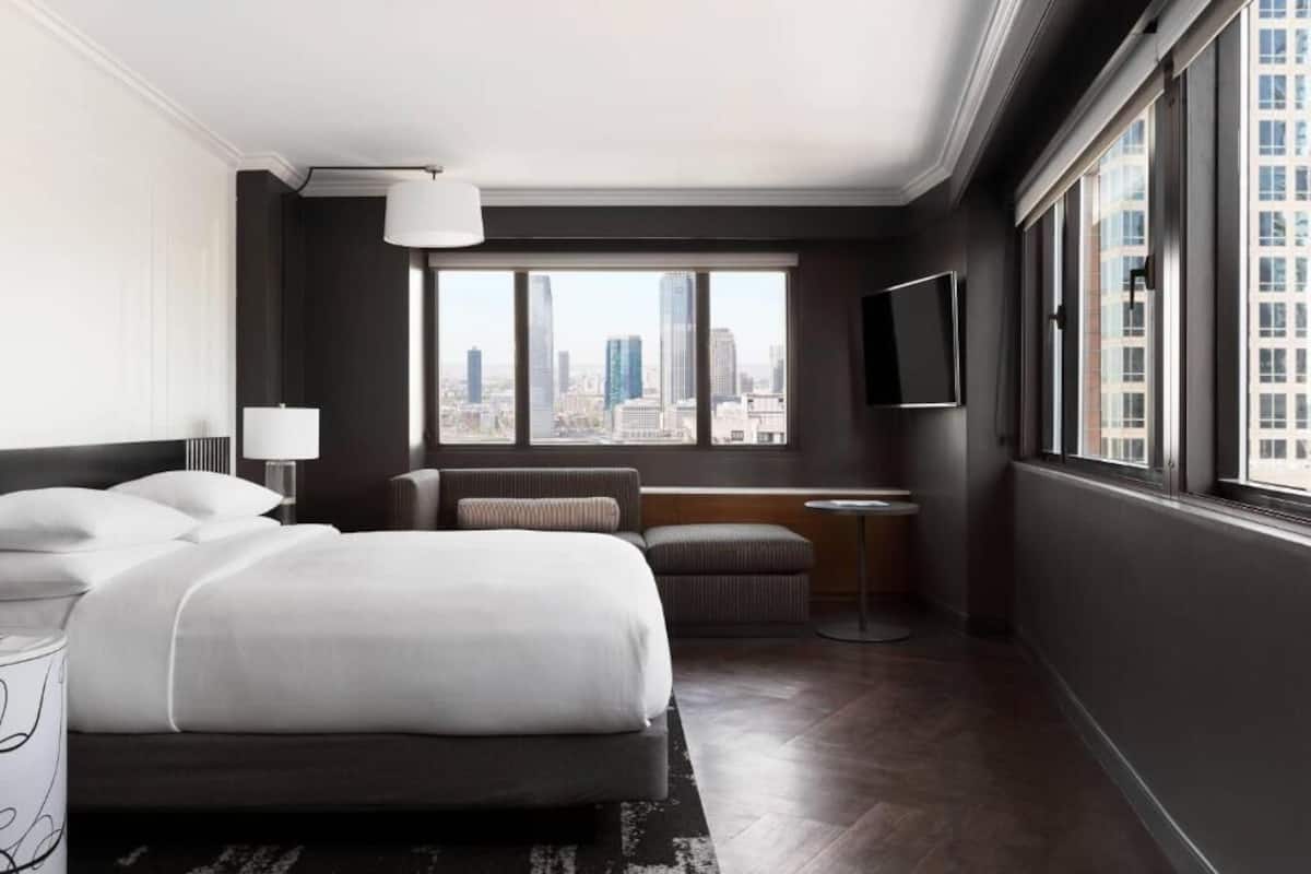 Una habitación de hotel moderna con una cama grande, zona de estar, televisor y vistas a la ciudad.