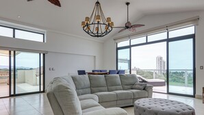 Un salon spacieux avec un canapé d’angle, un pouf rond, un ventilateur de plafond et de grandes fenêtres offrant une vue sur la ville.