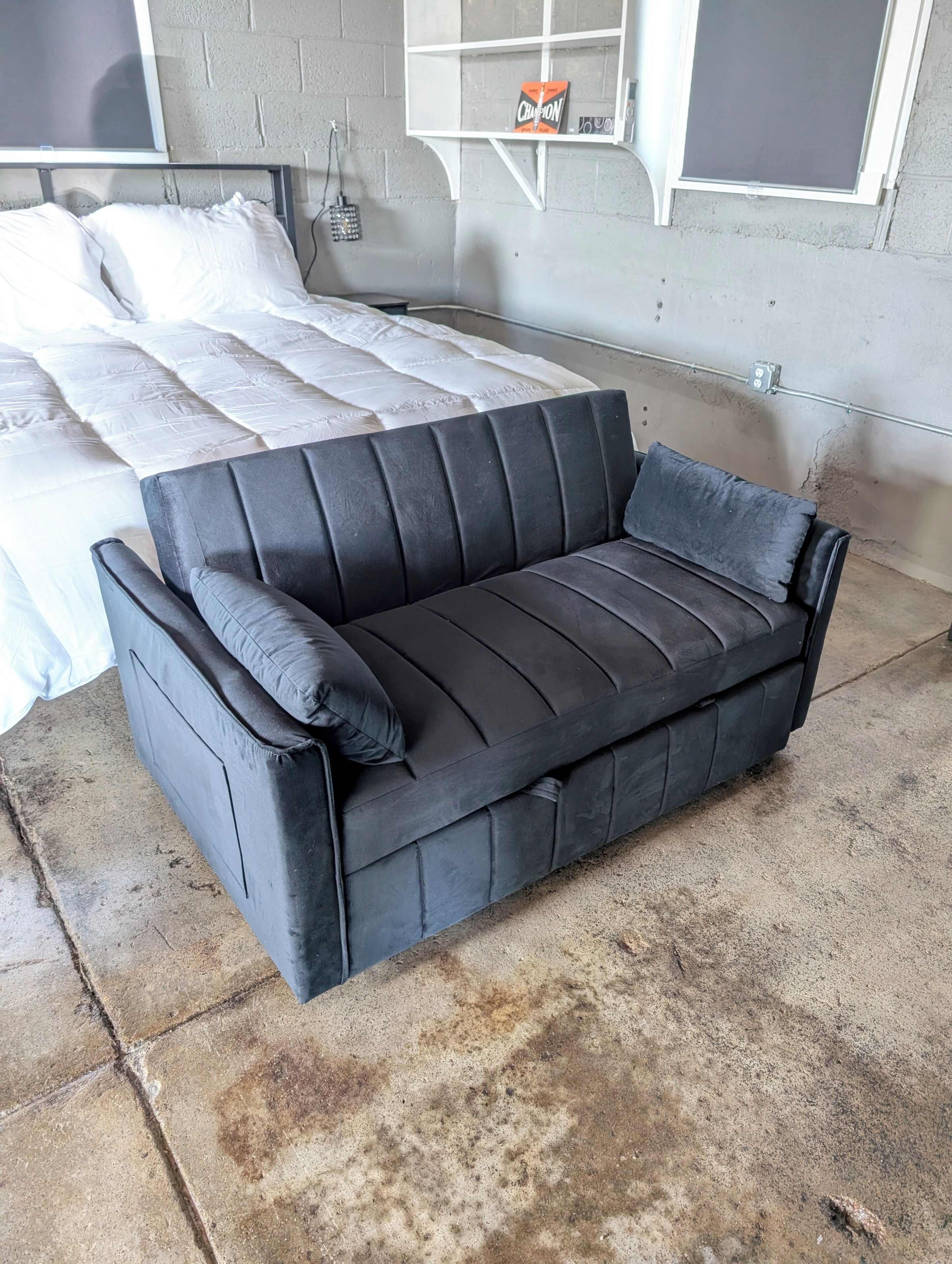 A black sofa with a cushion, a bed with white bedding, and a brick wall.