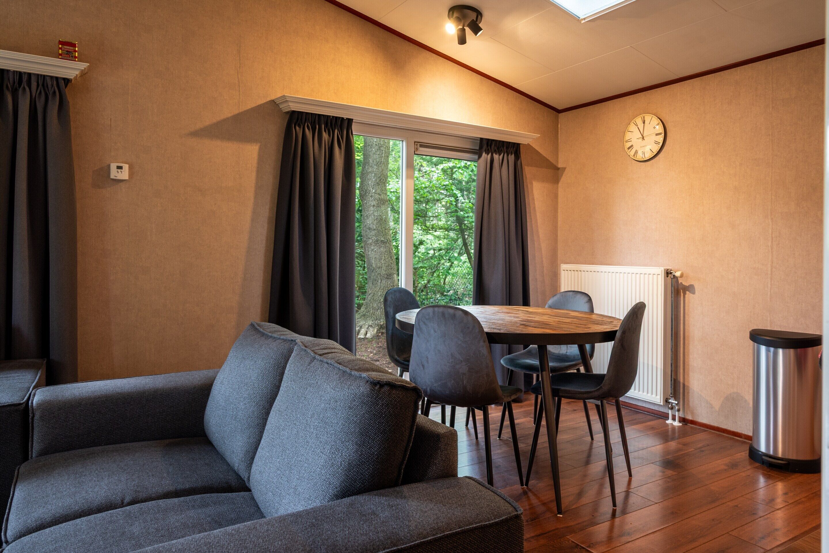 Une pièce confortable avec un canapé, une table à manger et des chaises, une horloge au mur et une fenêtre avec des rideaux.