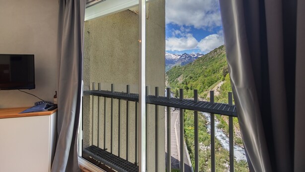 Una stanza con una finestra che si affaccia su un paesaggio montuoso, un tavolino con un televisore e una tenda.