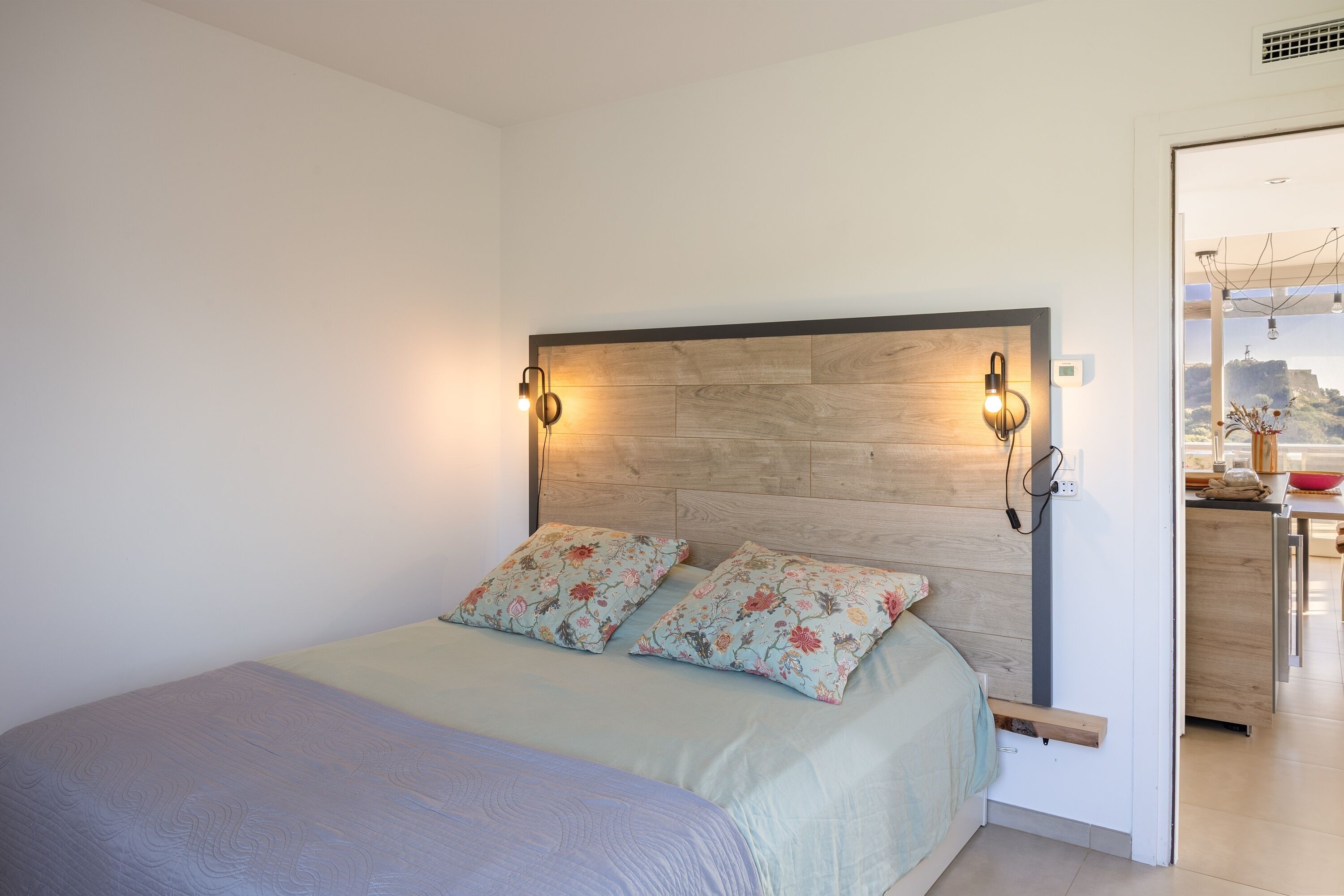 A bedroom with a bed, two wall-mounted lights, a wooden headboard, and a view of a kitchen area.
