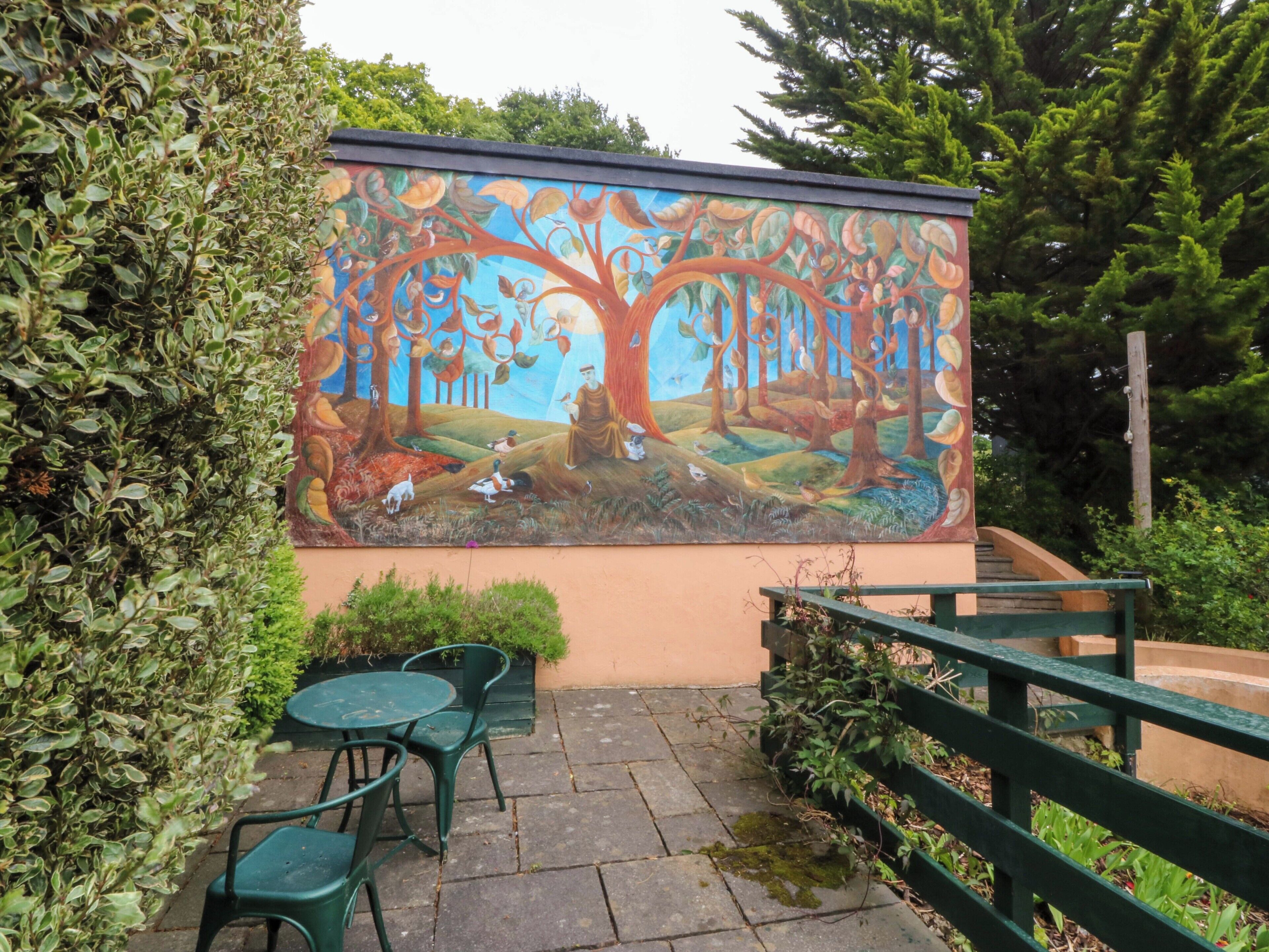 Un mural de un árbol con animales y una persona en una pared, mobiliario exterior y una valla de madera.