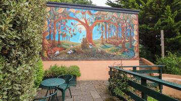 Un mural de un árbol con animales y una persona en una pared, mobiliario exterior y una valla de madera.