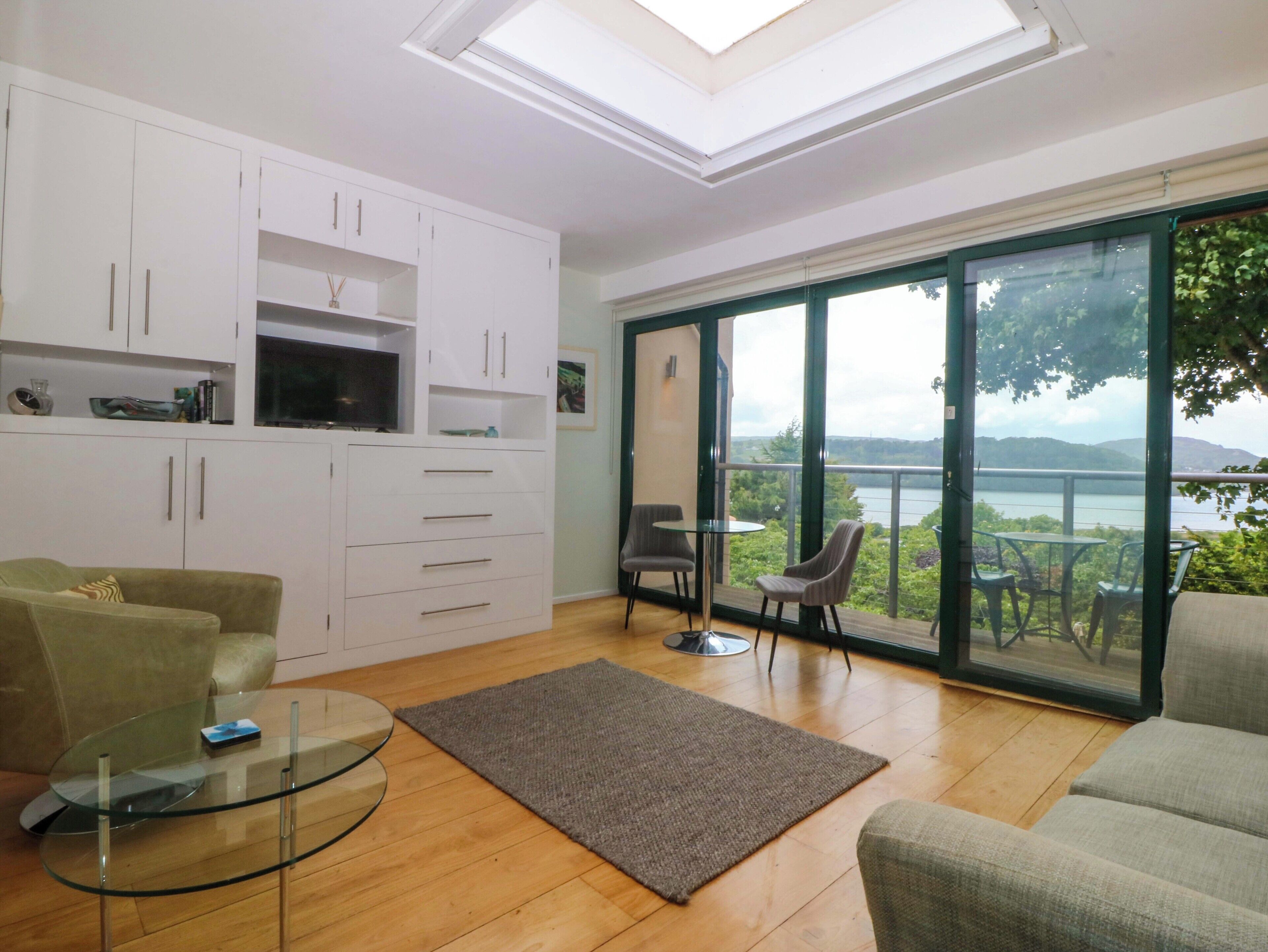 A modern living room with a glass table, a sofa, a TV, and a view of a lake through large glass doors.