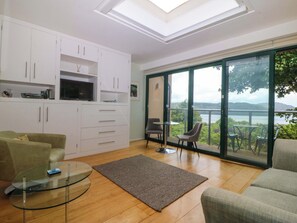 A modern living room with a glass table, a sofa, a TV, and a view of a lake through large glass doors. - The Treehouse (Colwyn Bay)