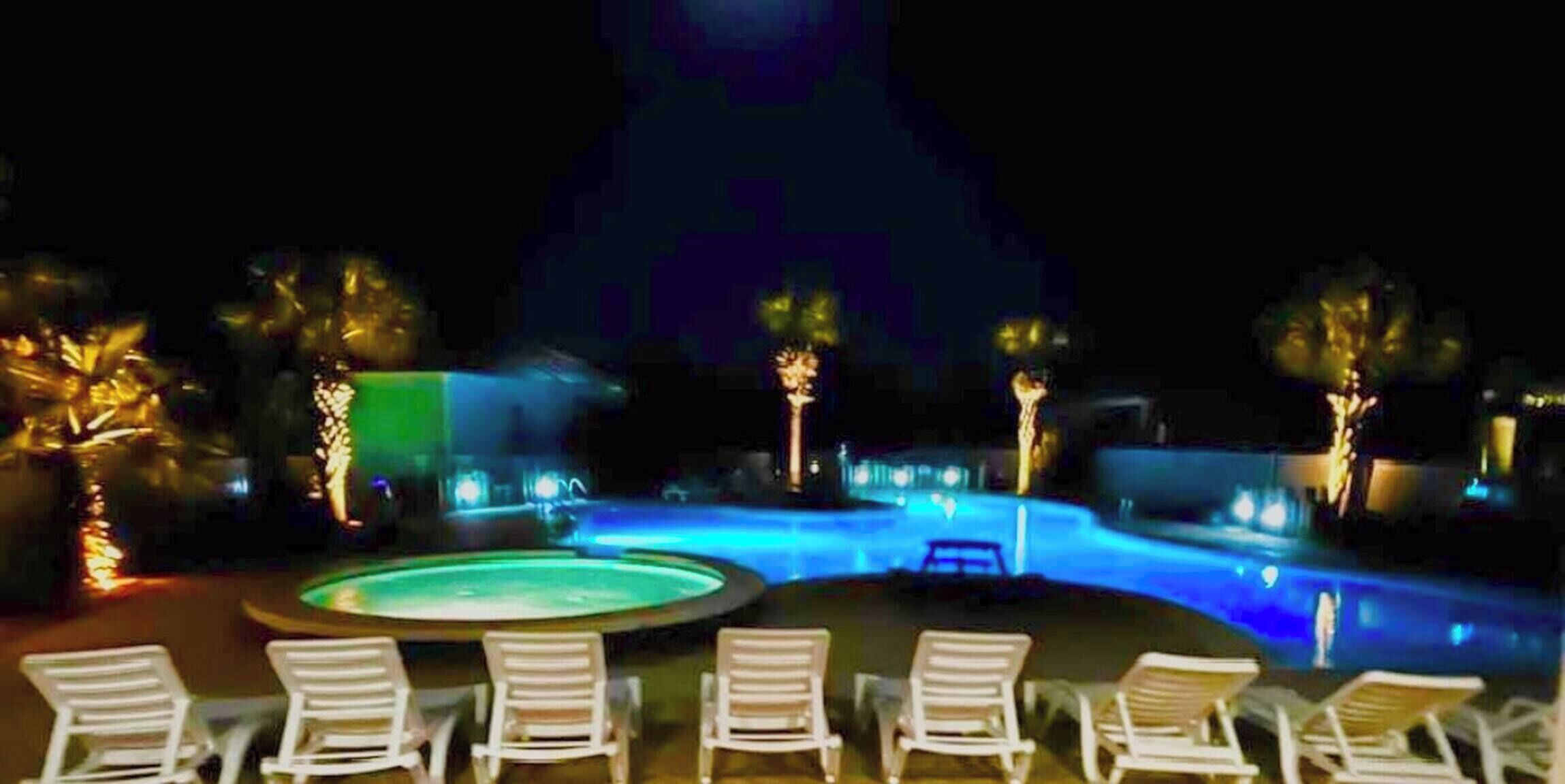 Ein Poolbereich bei Nacht mit Liegestühlen und beleuchteten Wasserspielen.